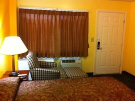 Una habitación de hotel con cama, ventana con cortinas, puerta y lámpara.