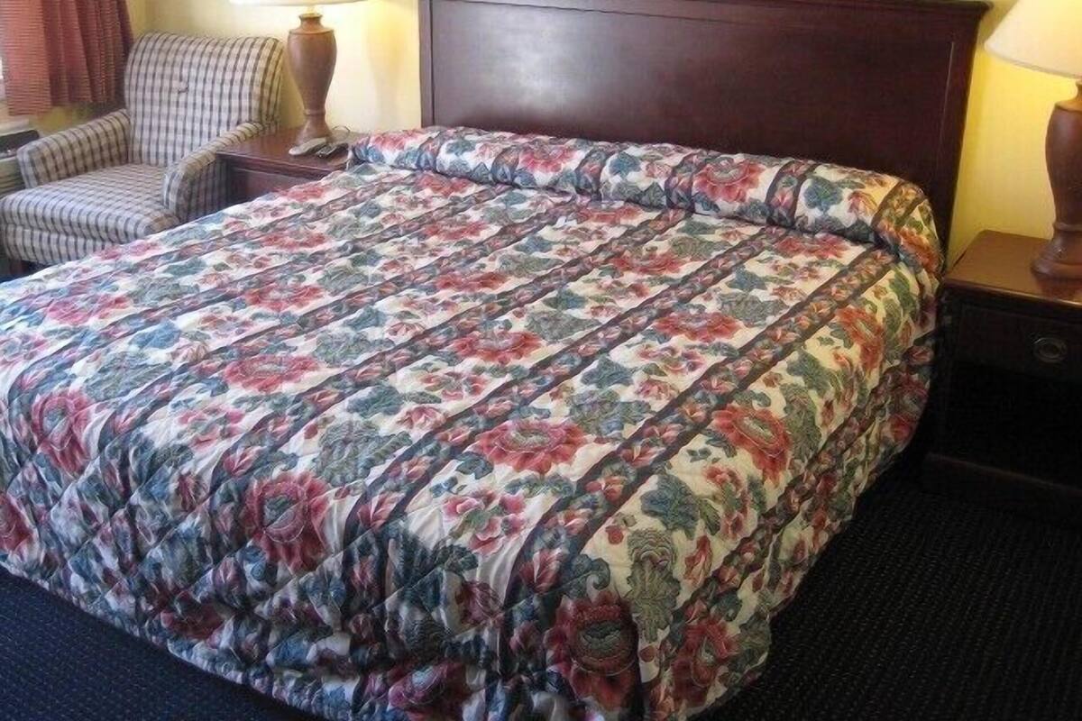 A neatly made bed with a floral comforter, a wooden headboard, a bedside table with a lamp, and a chair in the corner.