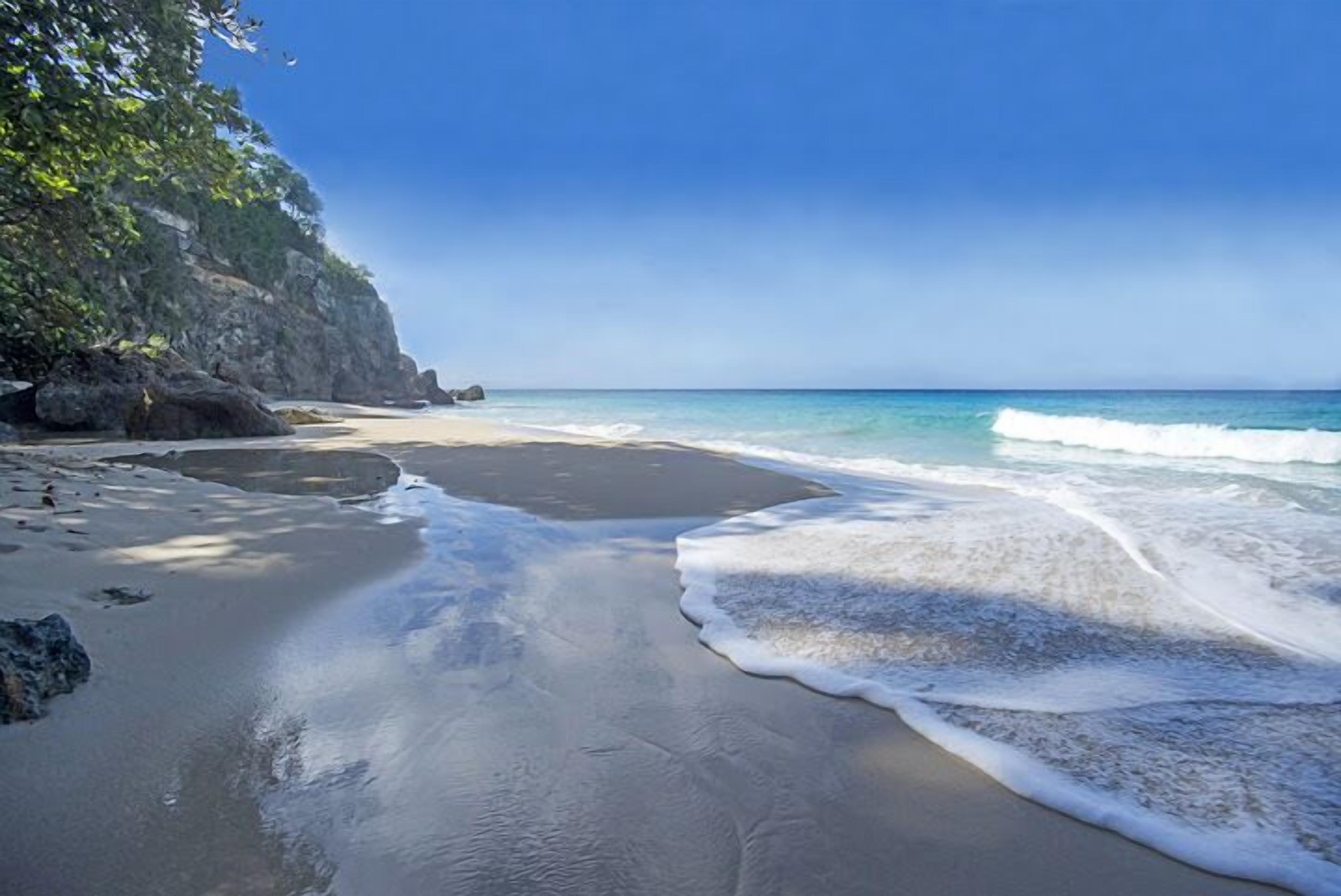 Une plage à l’eau d’un bleu limpide, avec une rive sablonneuse et une falaise rocheuse.