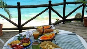 Une table dressée avec des aliments pour le petit-déjeuner, notamment des fruits, du pain et une boisson, sur une nappe à carreaux, donnant sur une plage.