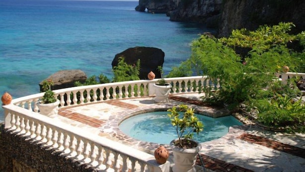 Un espace piscine doté d’une terrasse en pierre, entouré d’une balustrade blanche, offrant une vue sur une mer d’un bleu limpide et une falaise rocheuse.