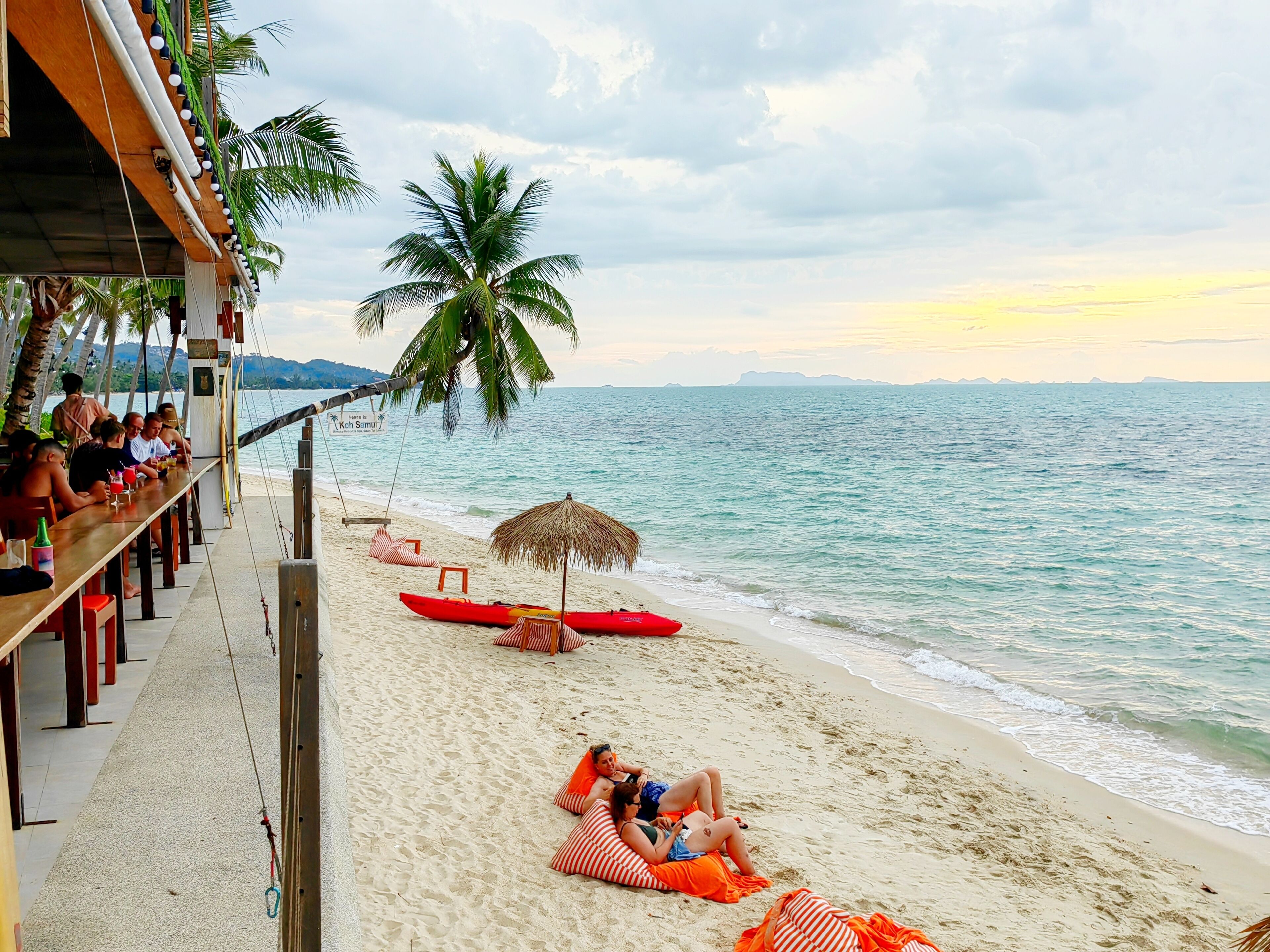 A beachside bar with a view of the ocean, a red canoe, and a couple relaxing on the sand.