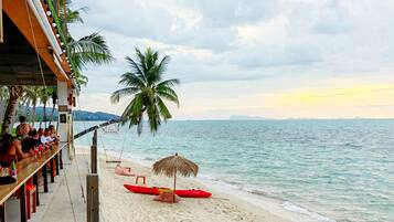 A beachside bar with a view of the ocean, a red canoe, and a couple relaxing on the sand.