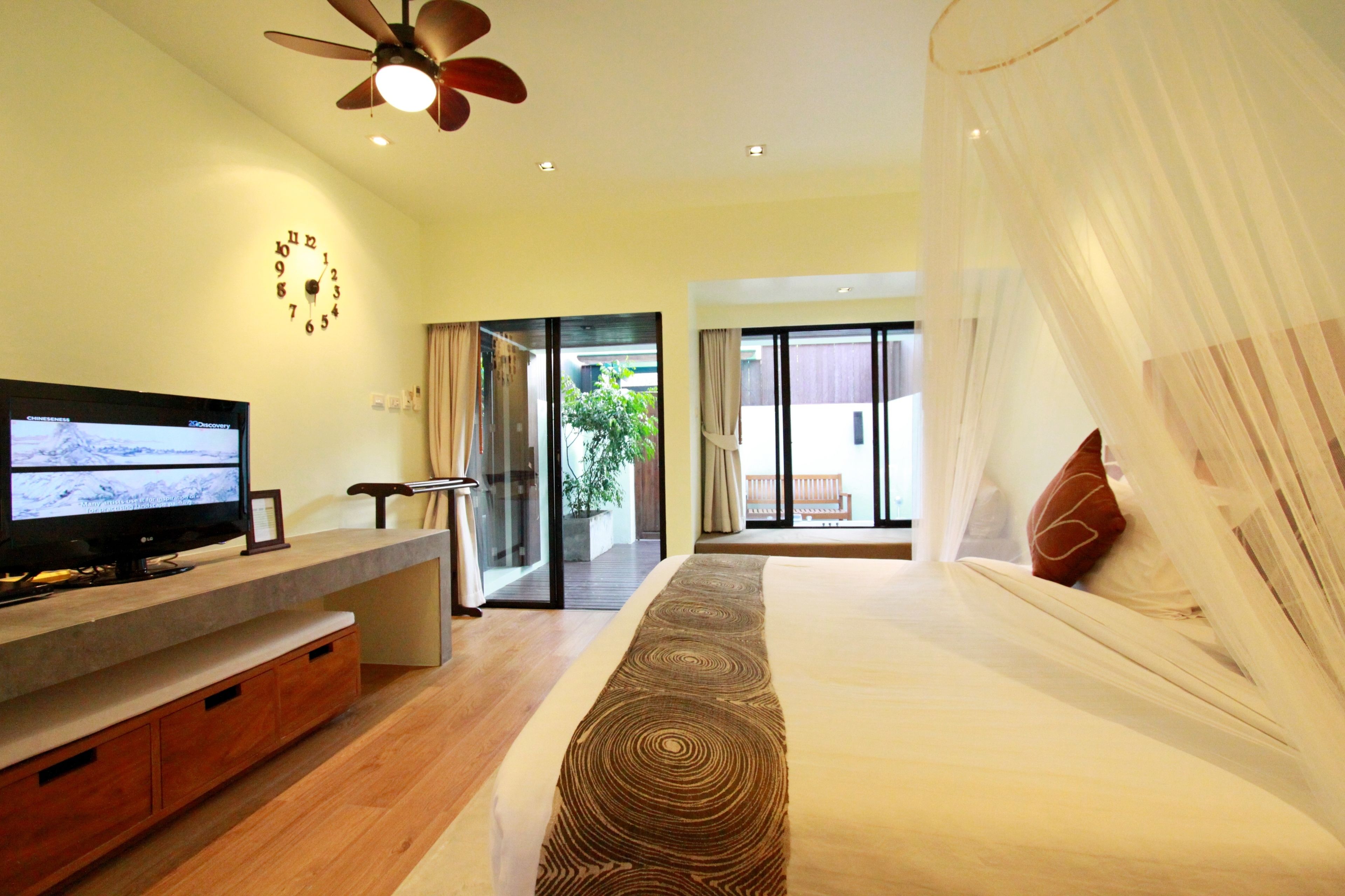 Un dormitorio con cama, televisión sobre un mueble de madera, ventilador de techo y vistas al exterior a través de una puerta de cristal.