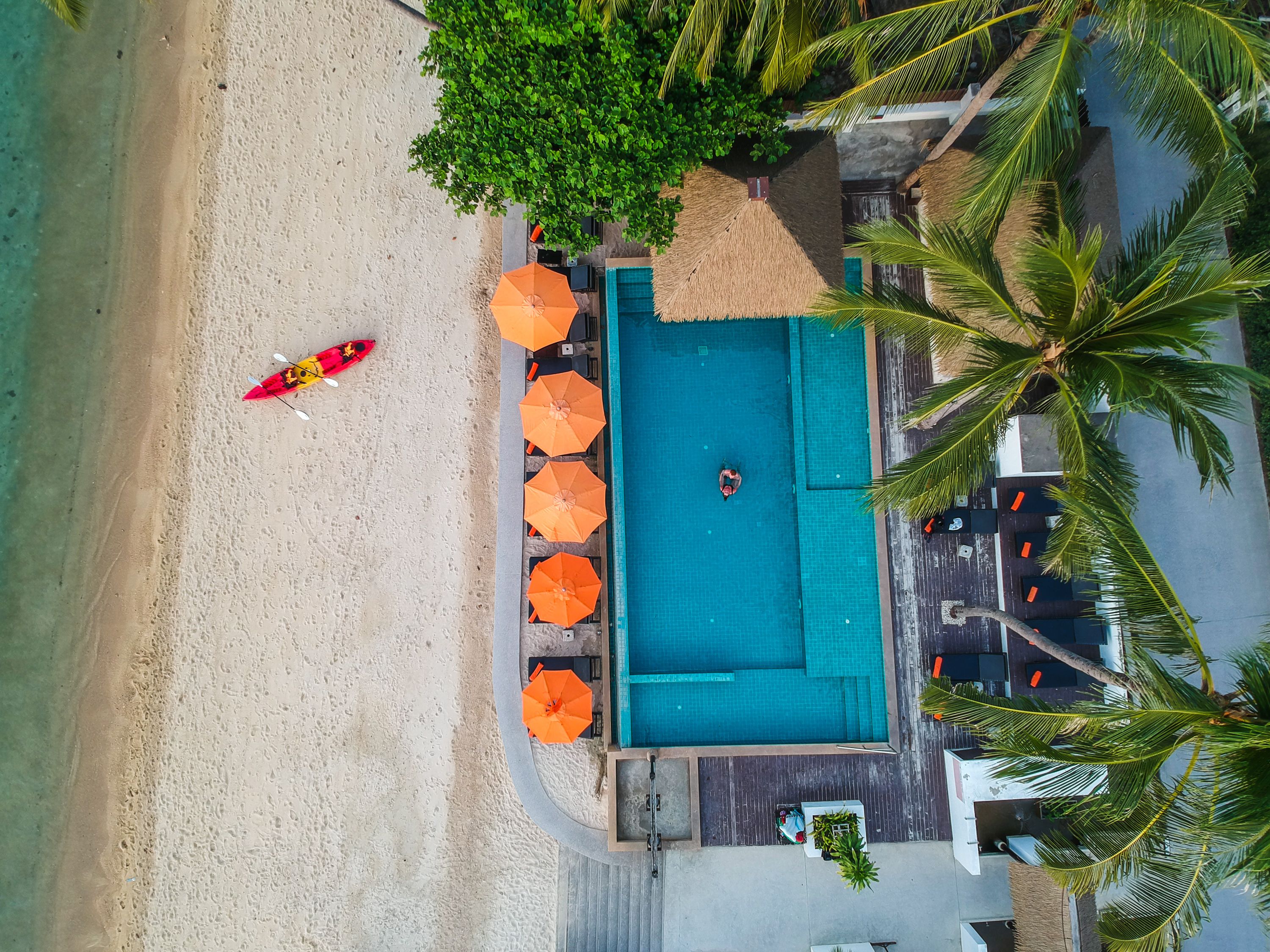 A pool area with a sandy beach, a red kayak, and orange umbrellas.