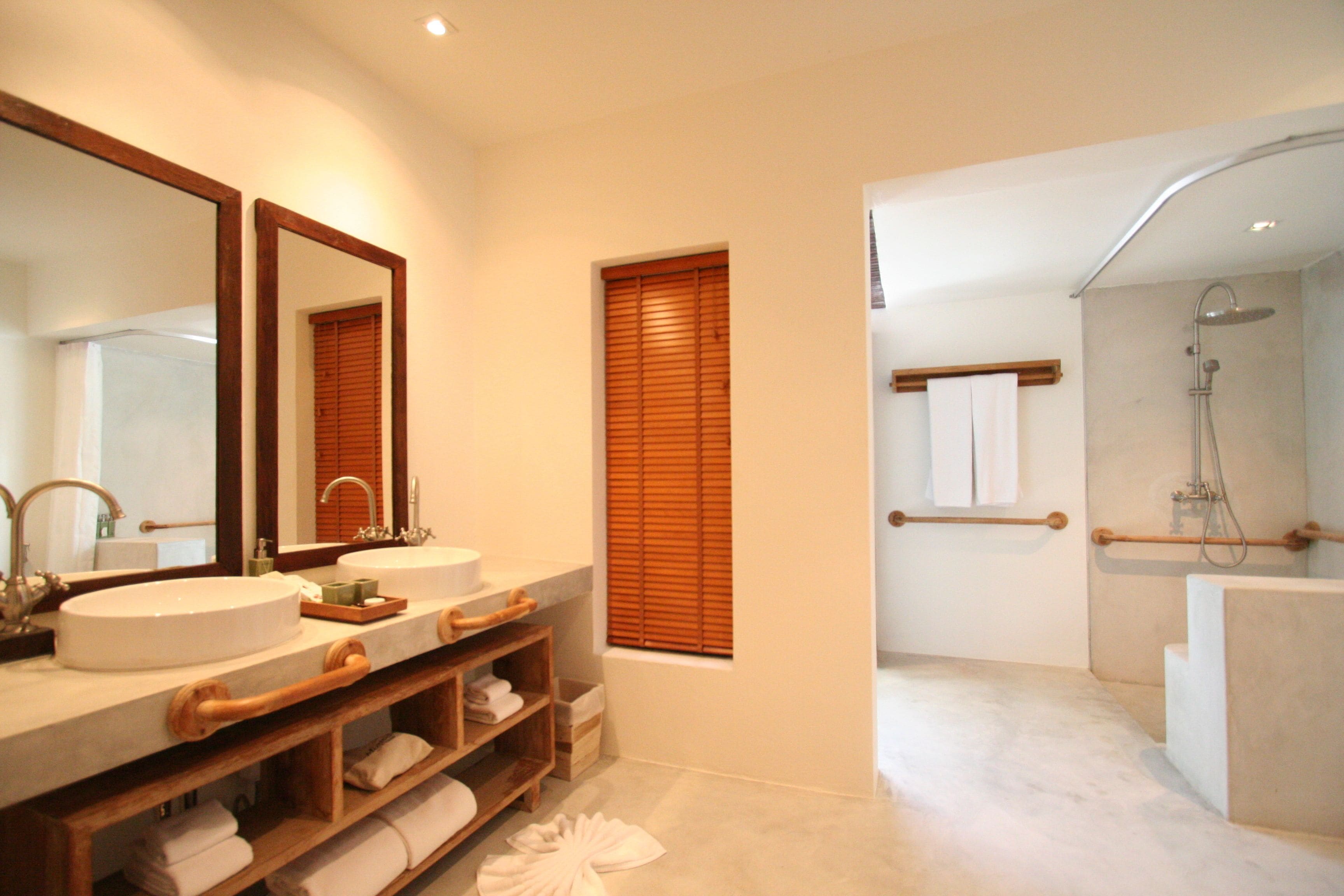 A modern bathroom with a large mirror, two sinks, wooden shelves with towels, a window with blinds, and a separate shower area.