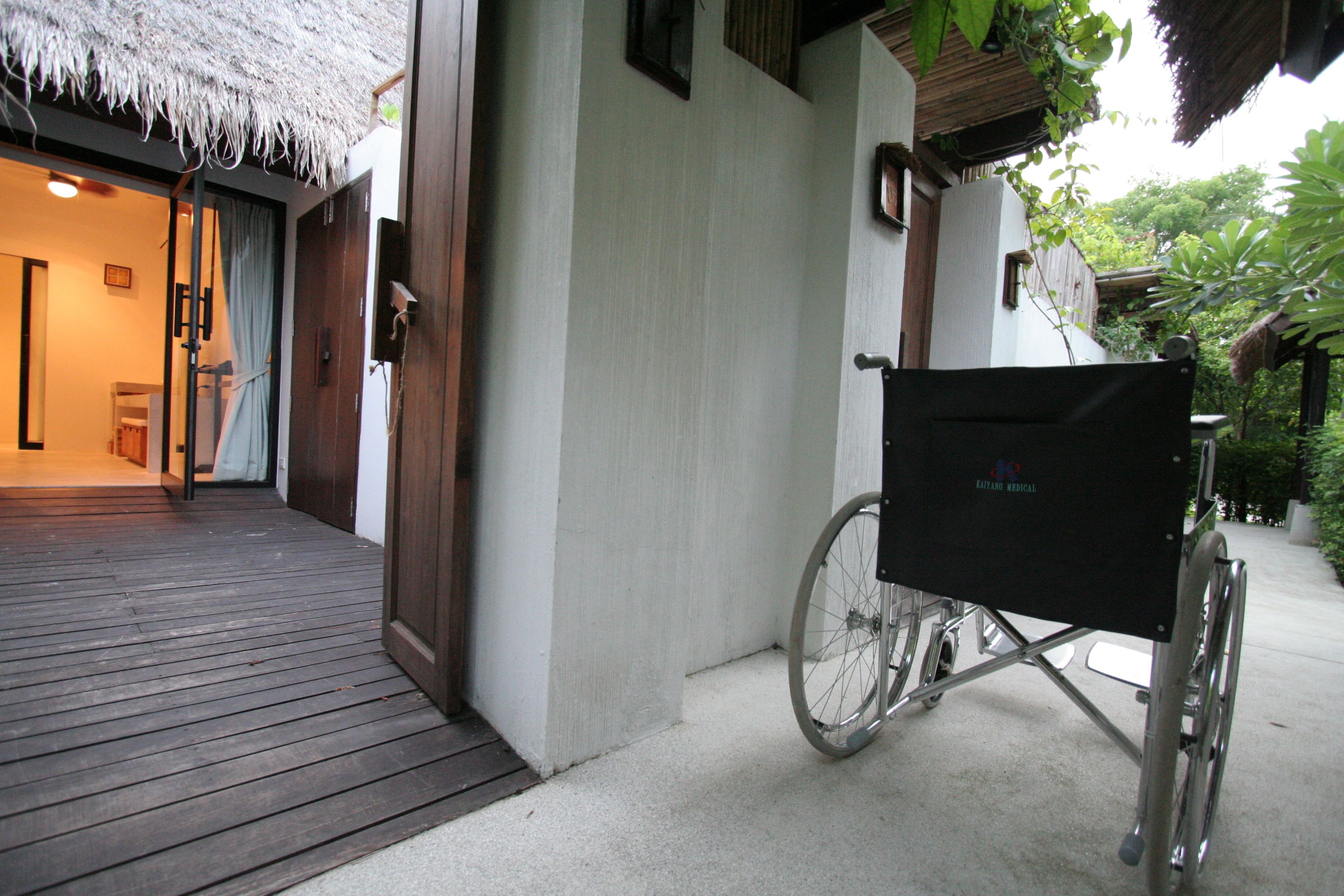 A wheelchair-accessible outdoor area with a wooden deck, a thatched roof, and a white building with a door.