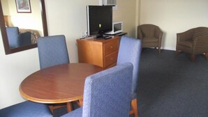 Una habitación con una mesa redonda de madera, dos sillas azules, un aparador de madera con un televisor y un microondas encima, y dos sillones a cuadros.