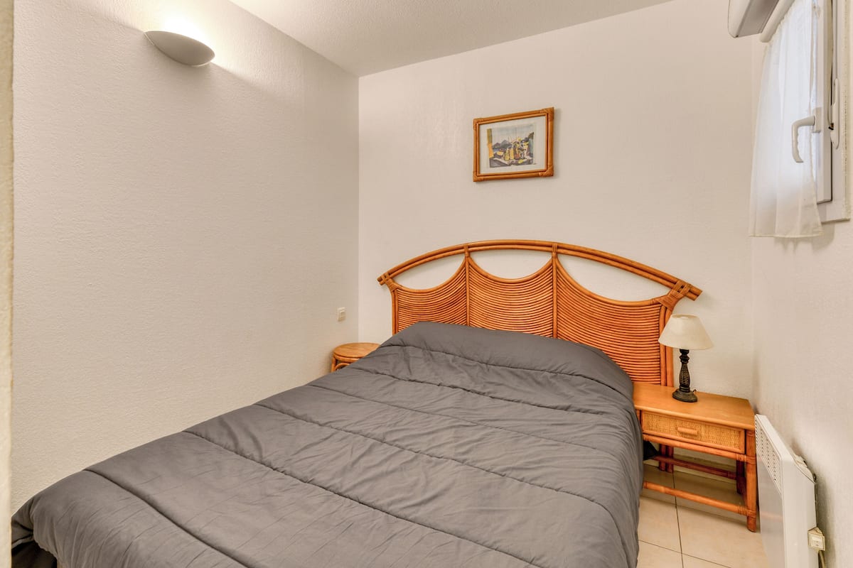 Ein Schlafzimmer mit einem Bett, einem Nachttisch, einer Lampe, einem gerahmten Bild an der Wand und einem Fenster mit Vorhängen.