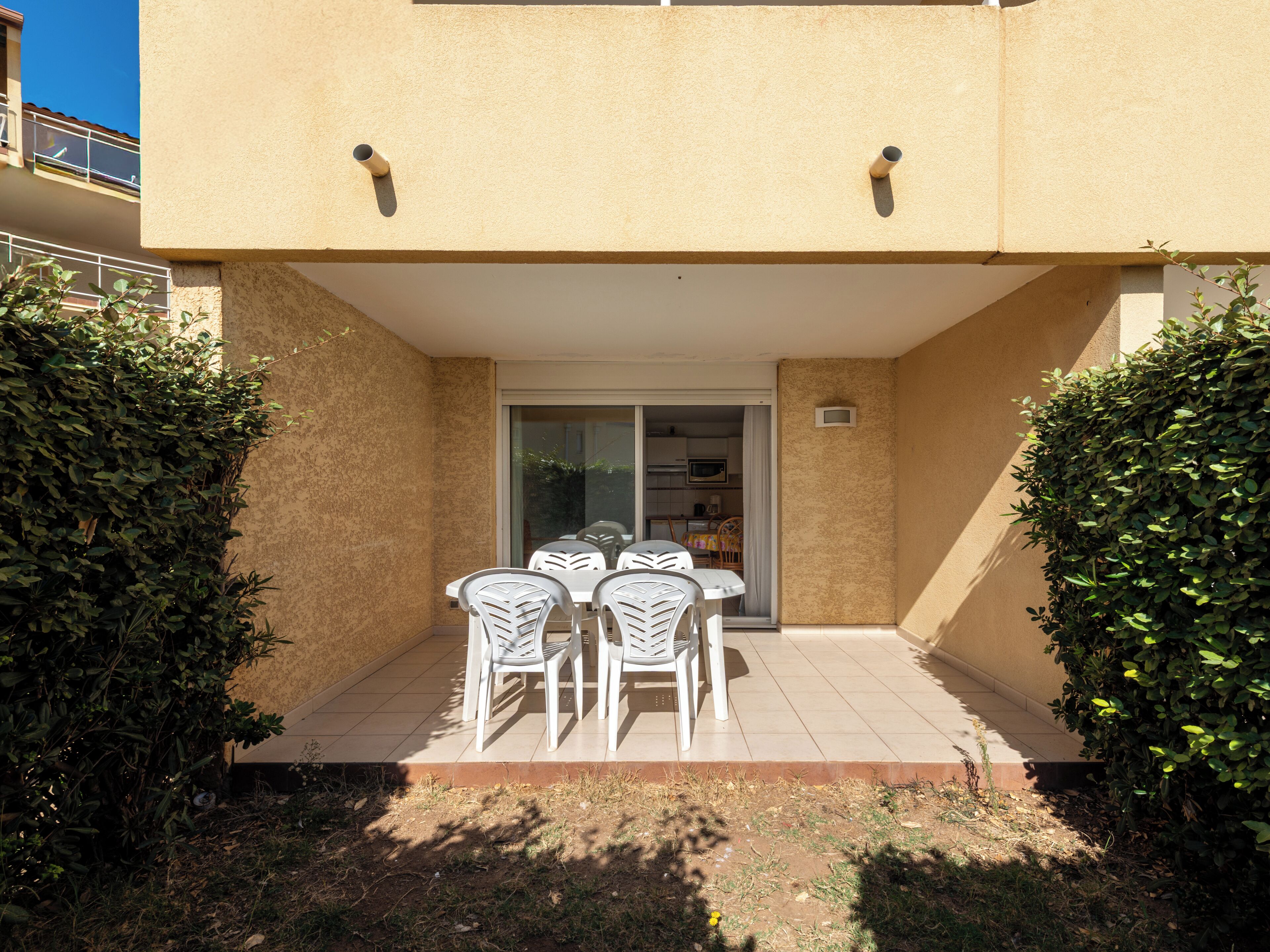 Un patio con una mesa y sillas, rodeado de setos, que conduce a una puerta de vidrio.