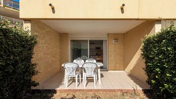 Un patio con una mesa y sillas, rodeado de setos, que conduce a una puerta de vidrio.