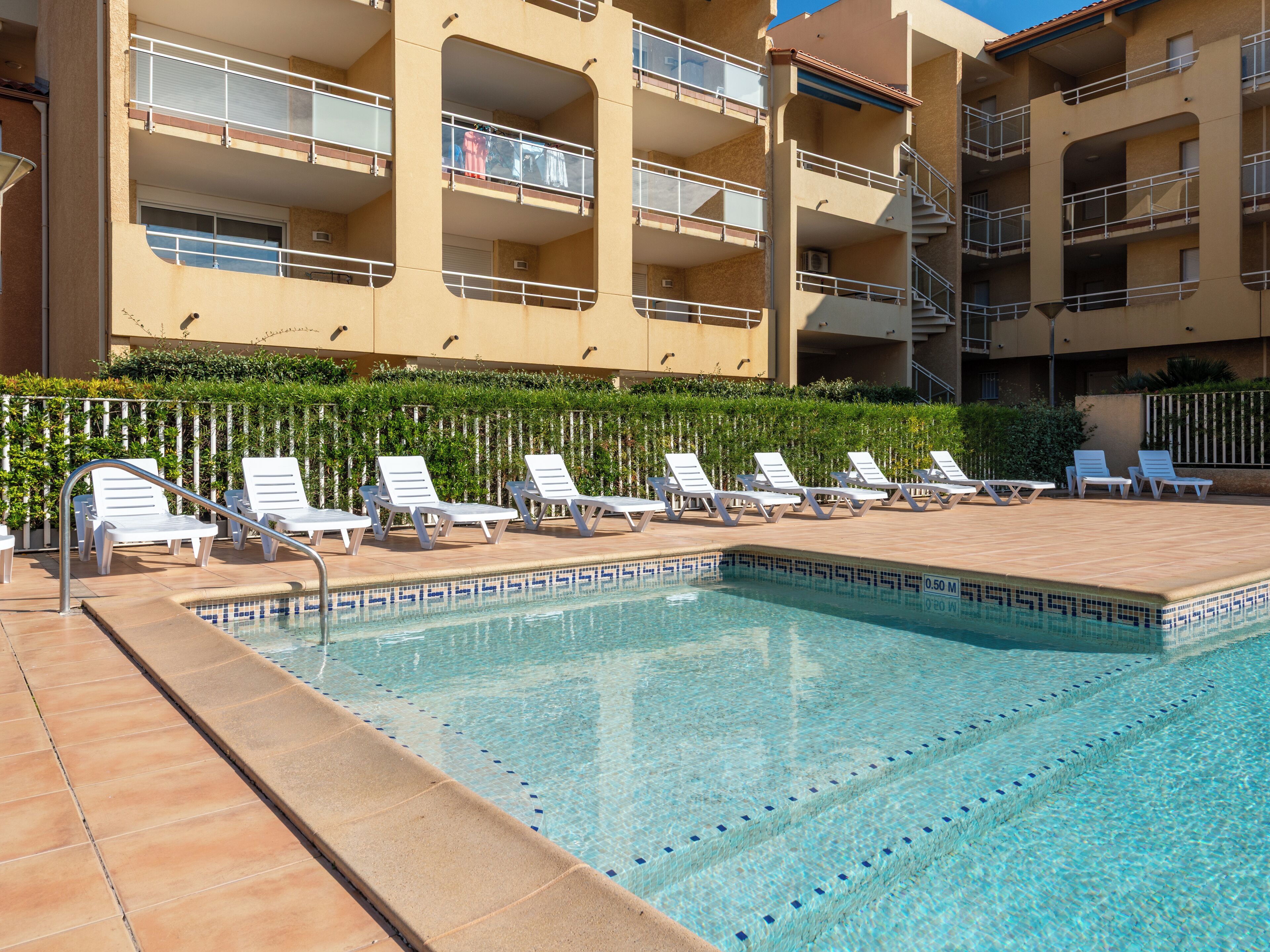 Une piscine entourée de chaises longues, une terrasse carrelée et, au fond, un immeuble à plusieurs étages doté de balcons.