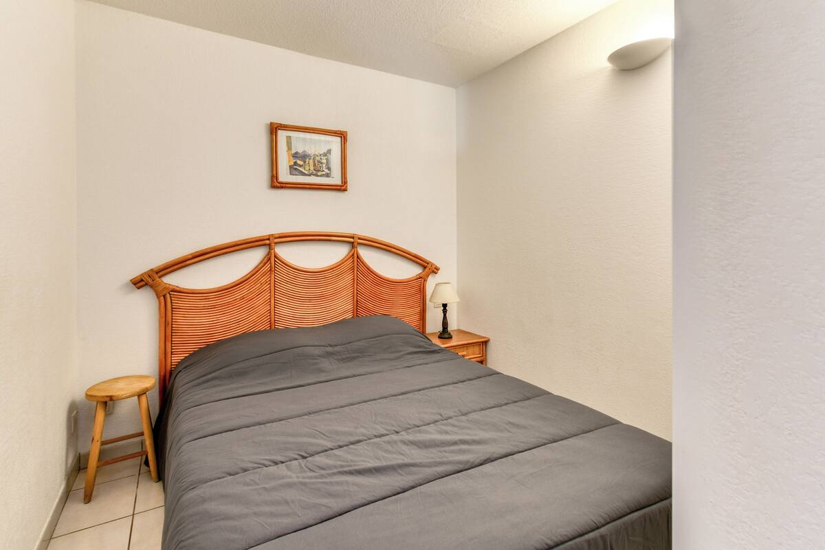 Ein ordentlich bezogenes Bett mit einem Holzkopfstück, ein kleiner Holzschemel, ein Nachttisch mit einer Lampe und ein gerahmtes Bild an der Wand.