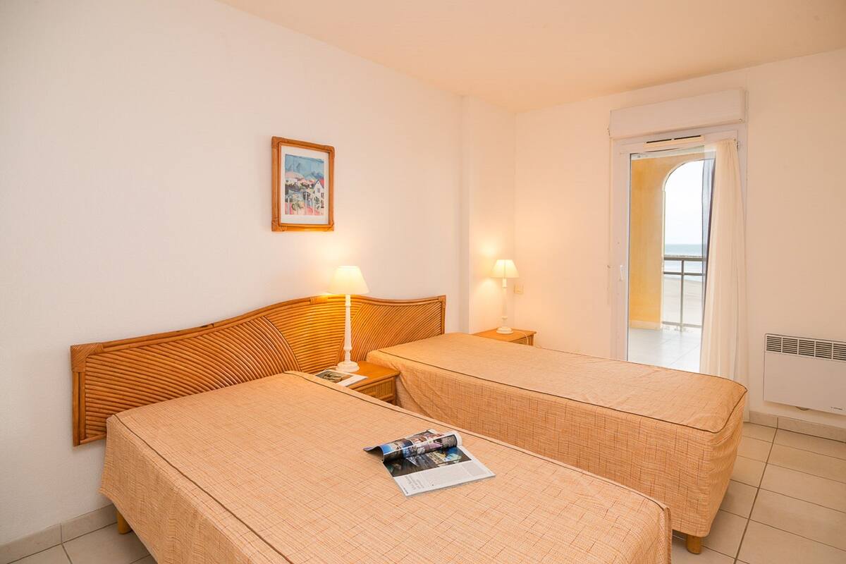 Ein Hotelzimmer mit zwei Einzelbetten, einem Nachttisch mit Lampe, einem gerahmten Bild an der Wand und einem Fenster mit Vorhängen.
