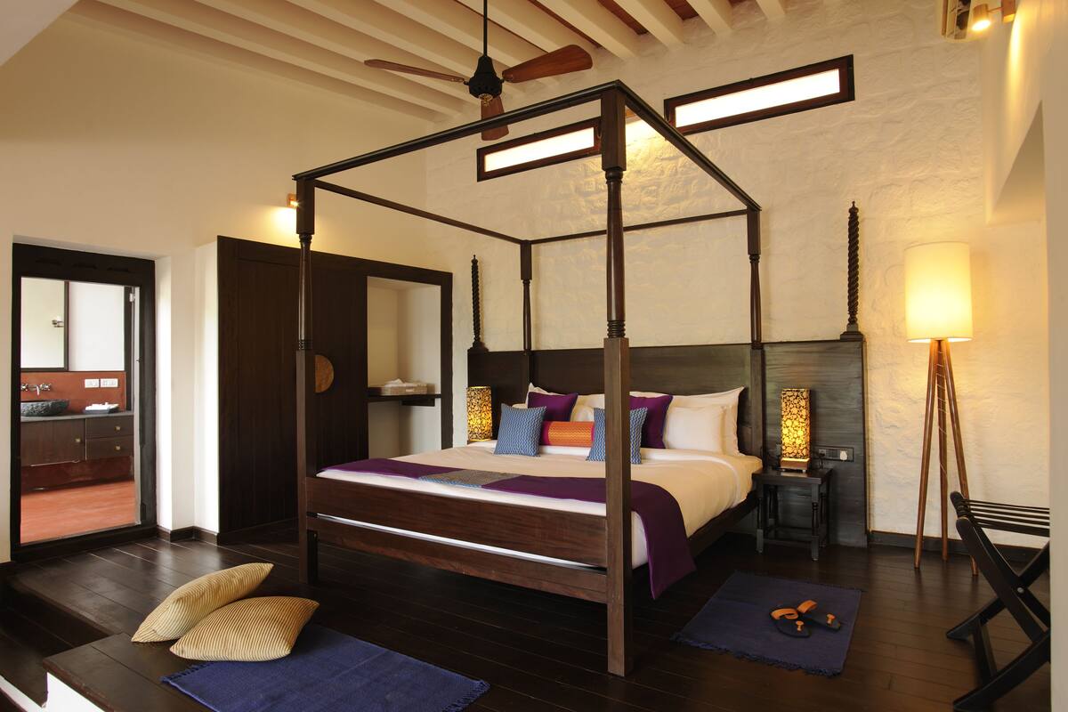 Un lit à baldaquin avec un cadre en bois, une table de chevet avec une lampe et un banc avec un coussin.