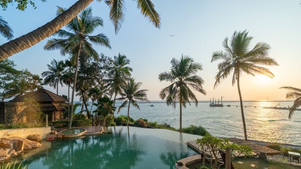 Piscina con vista al mar, rodeada de palmeras y un edificio con techo de paja.