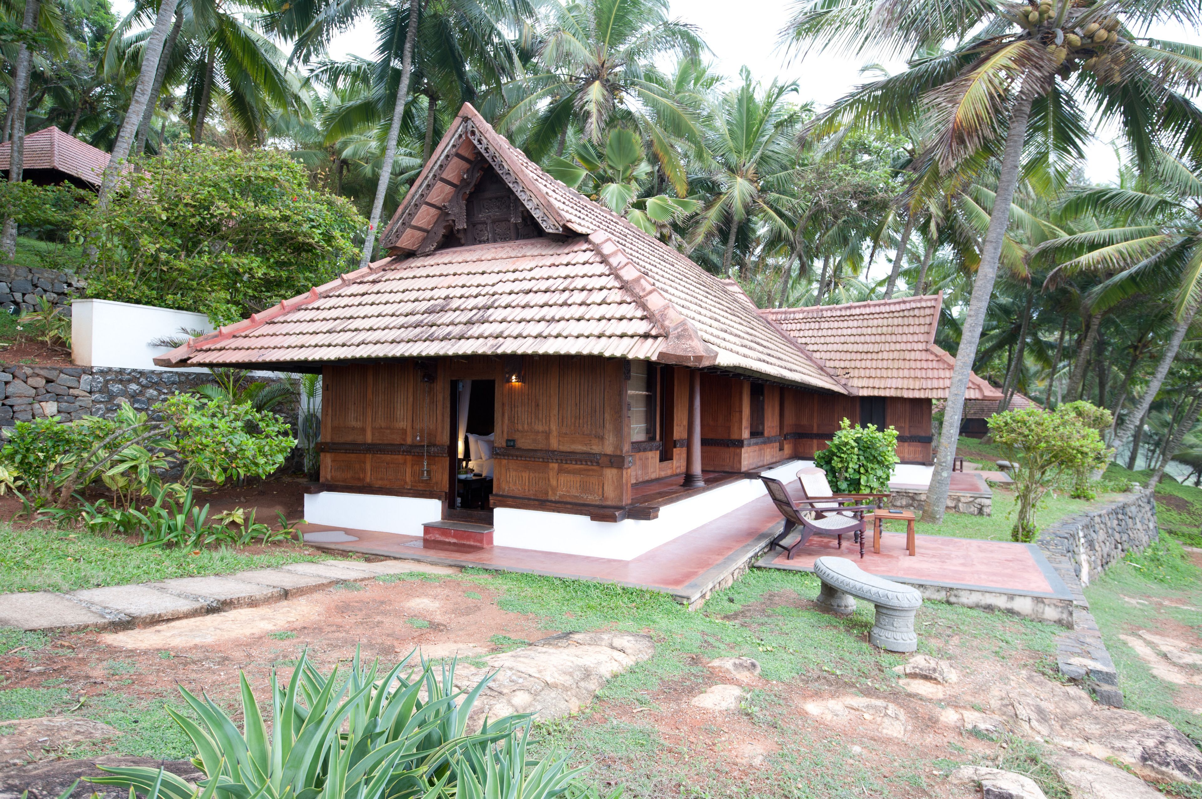 A traditional wooden house with a tiled roof, surrounded by lush greenery and palm trees.