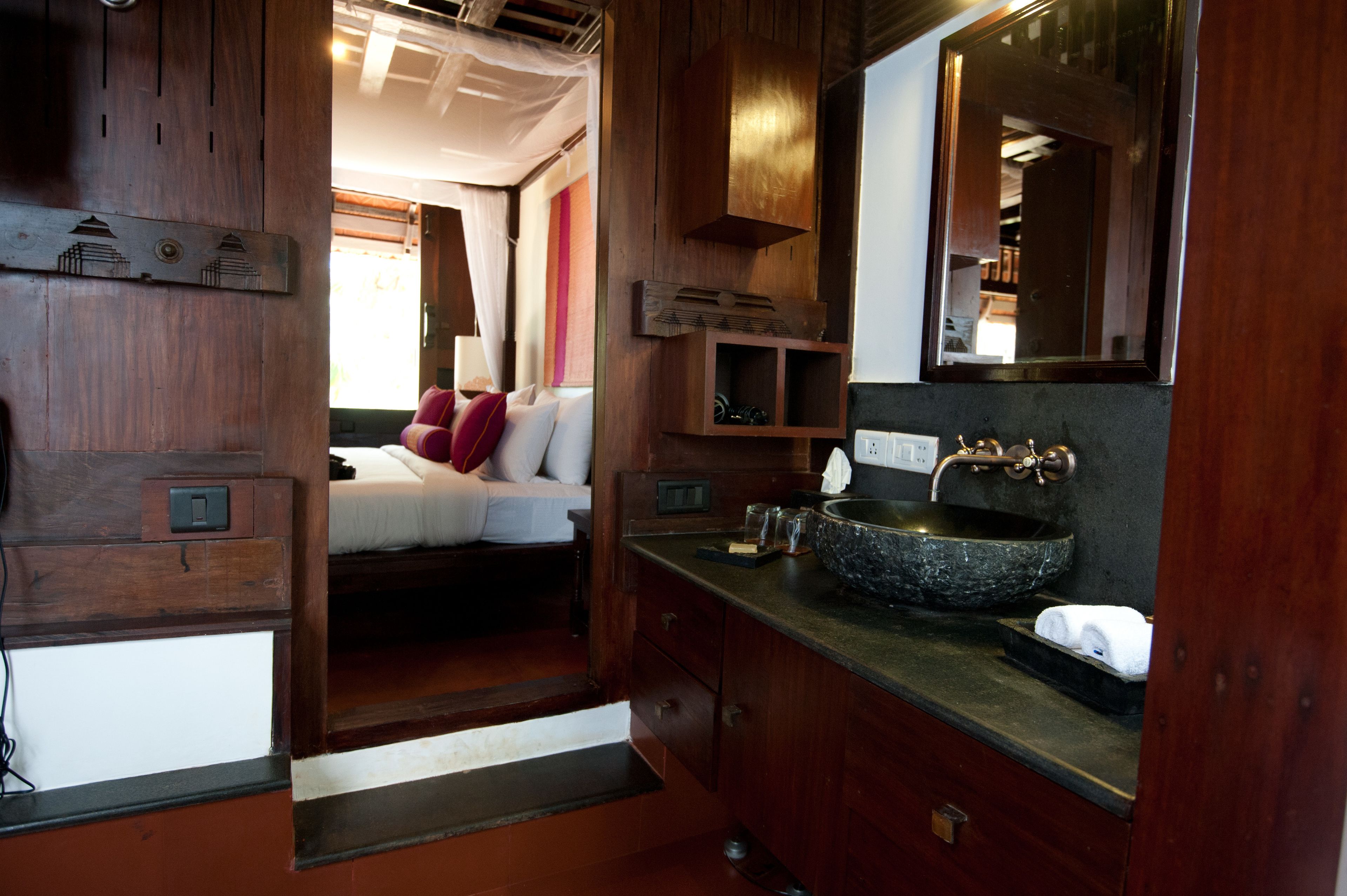 A compact, wood-finished bathroom with a built-in sink, a mirror, and a bed with red pillows.