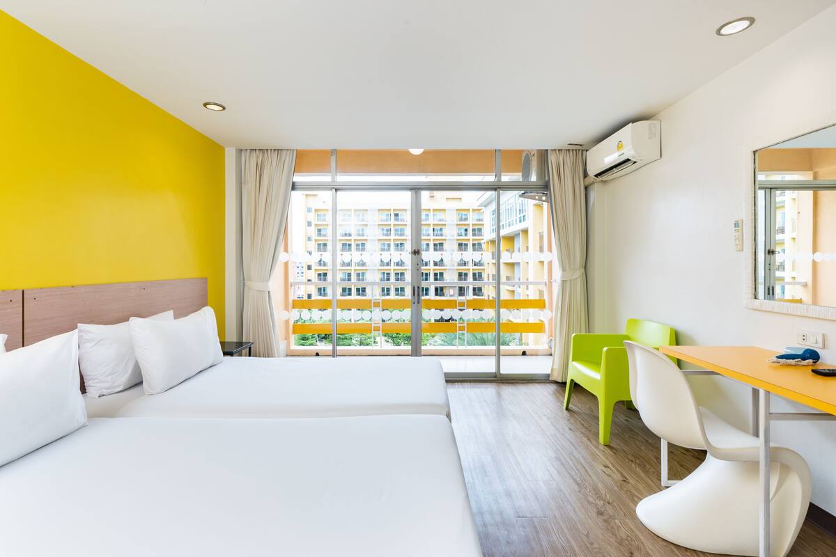 Quarto de hotel com uma cama, escrivaninha, cadeira e uma janela grande com vista para um edifício.
