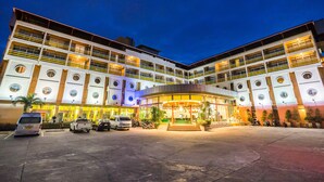 A multi-story hotel with a central atrium, parking area, and several vehicles parked in front. - Bella Express (Pattaya)