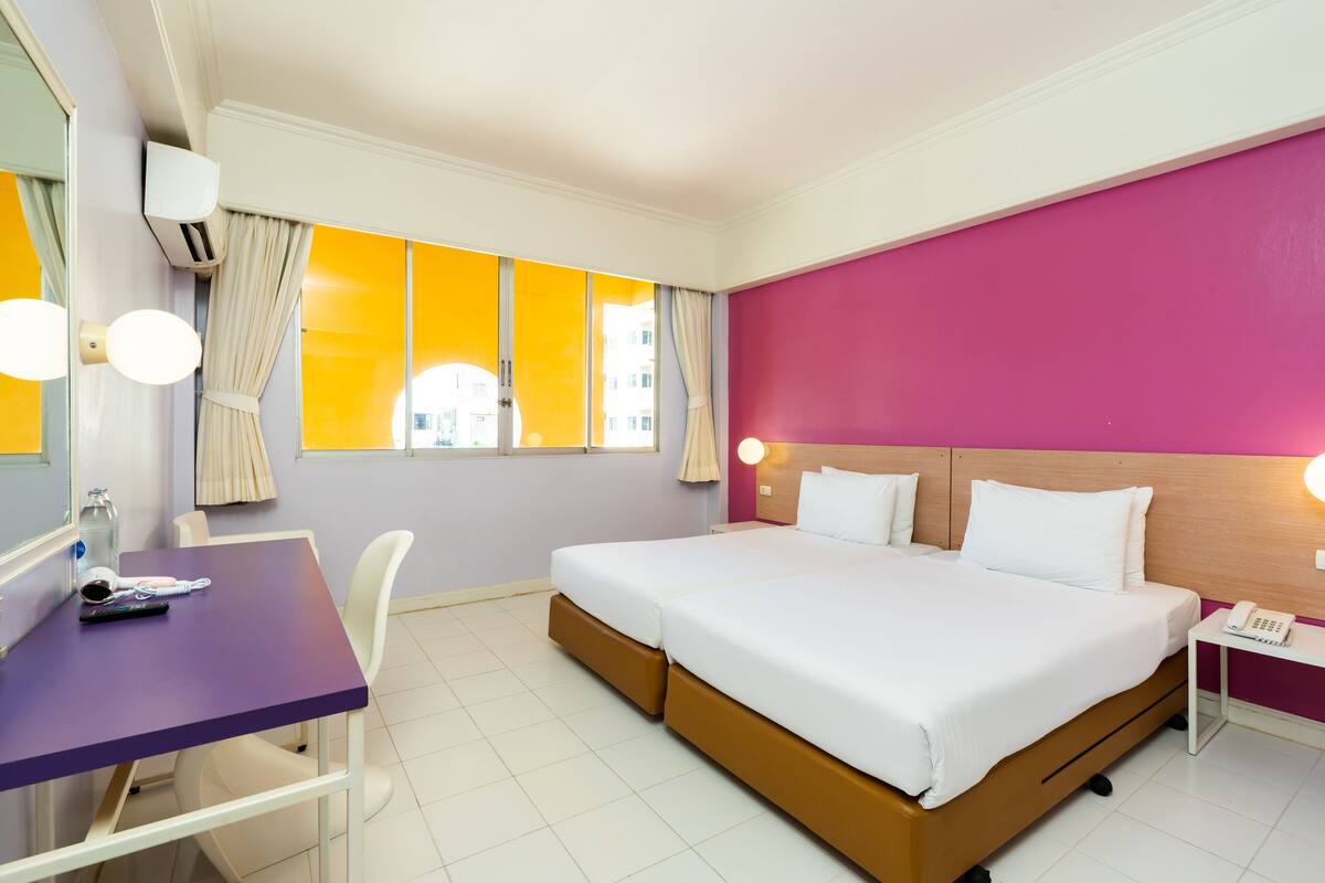 Quarto de hotel com uma cama grande, uma escrivaninha com cadeira, mesa de cabeceira com telefone e janela com cortinas.