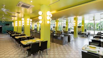 A brightly lit dining area with yellow walls, multiple ceiling fans, and round hanging lights. Tables are set with plates and cutlery, and there are chairs arranged around them.