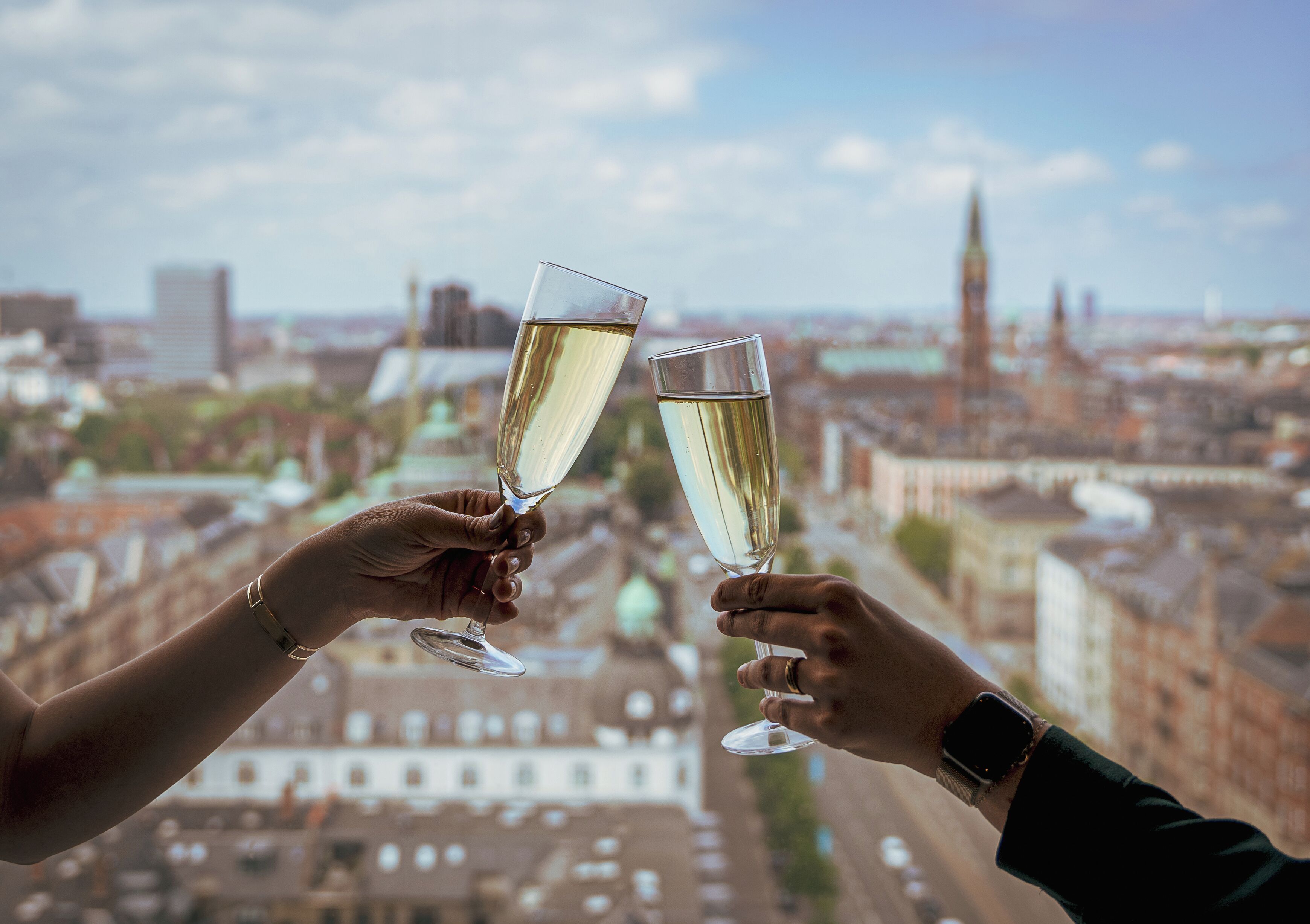 Two hands holding champagne glasses with a cityscape in the background.