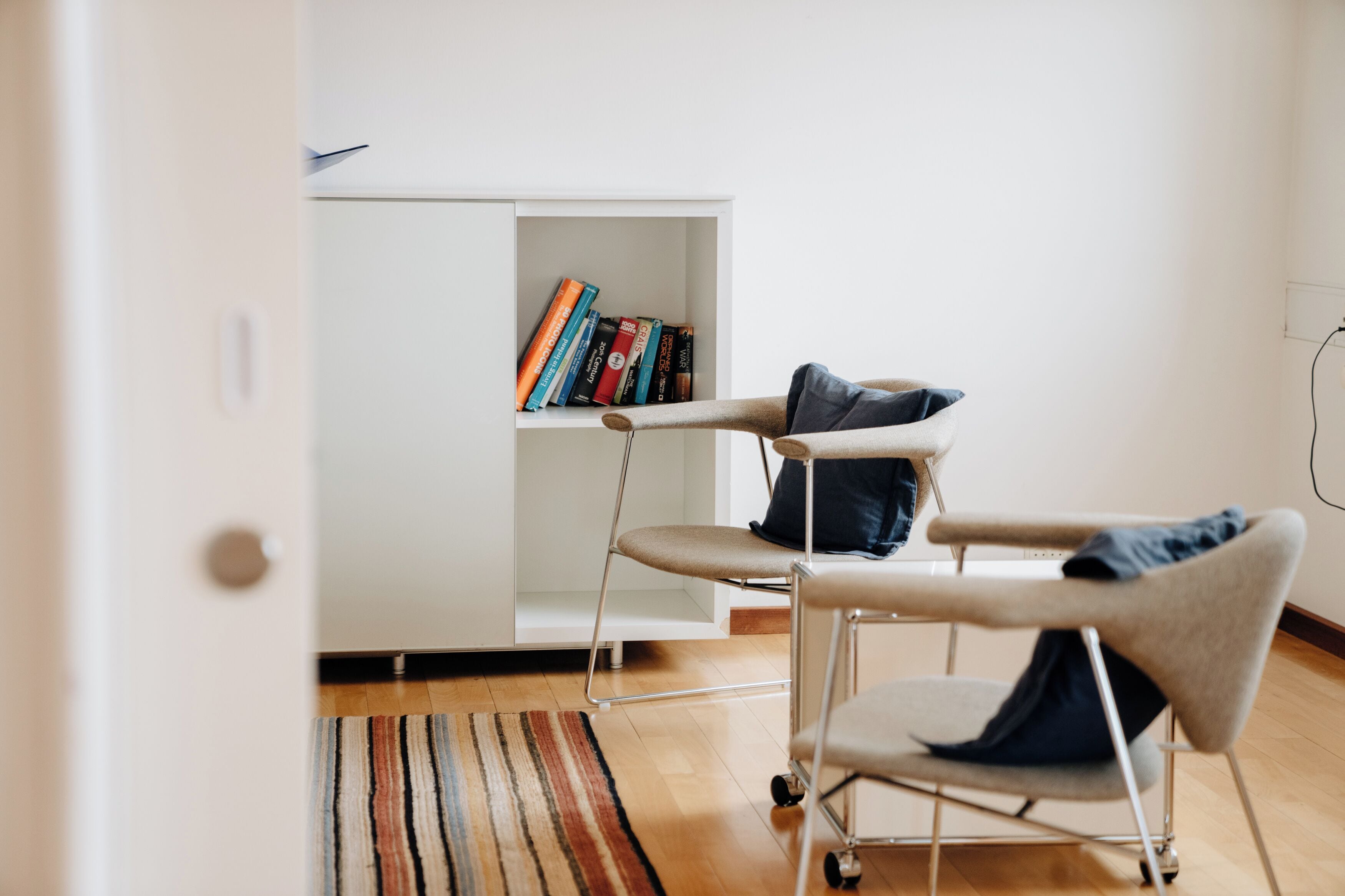 A modern room with a white cabinet, two chairs with cushions, a striped rug, and a bookshelf with books.