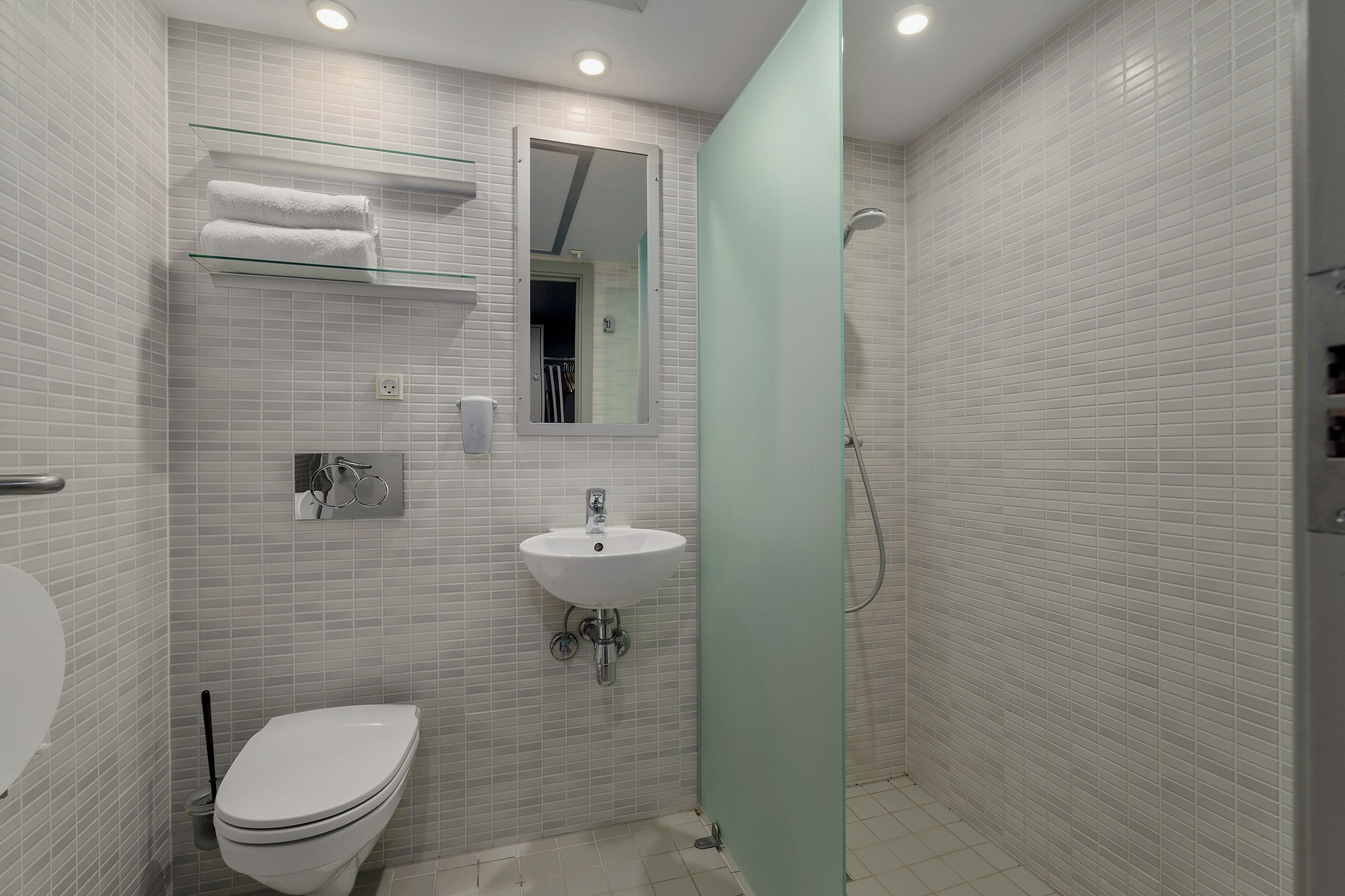 Ein Badezimmer mit weiß gefliesten Wänden, einem weißen Waschbecken, einer Toilette und einem Glastischchen mit Handtüchern.