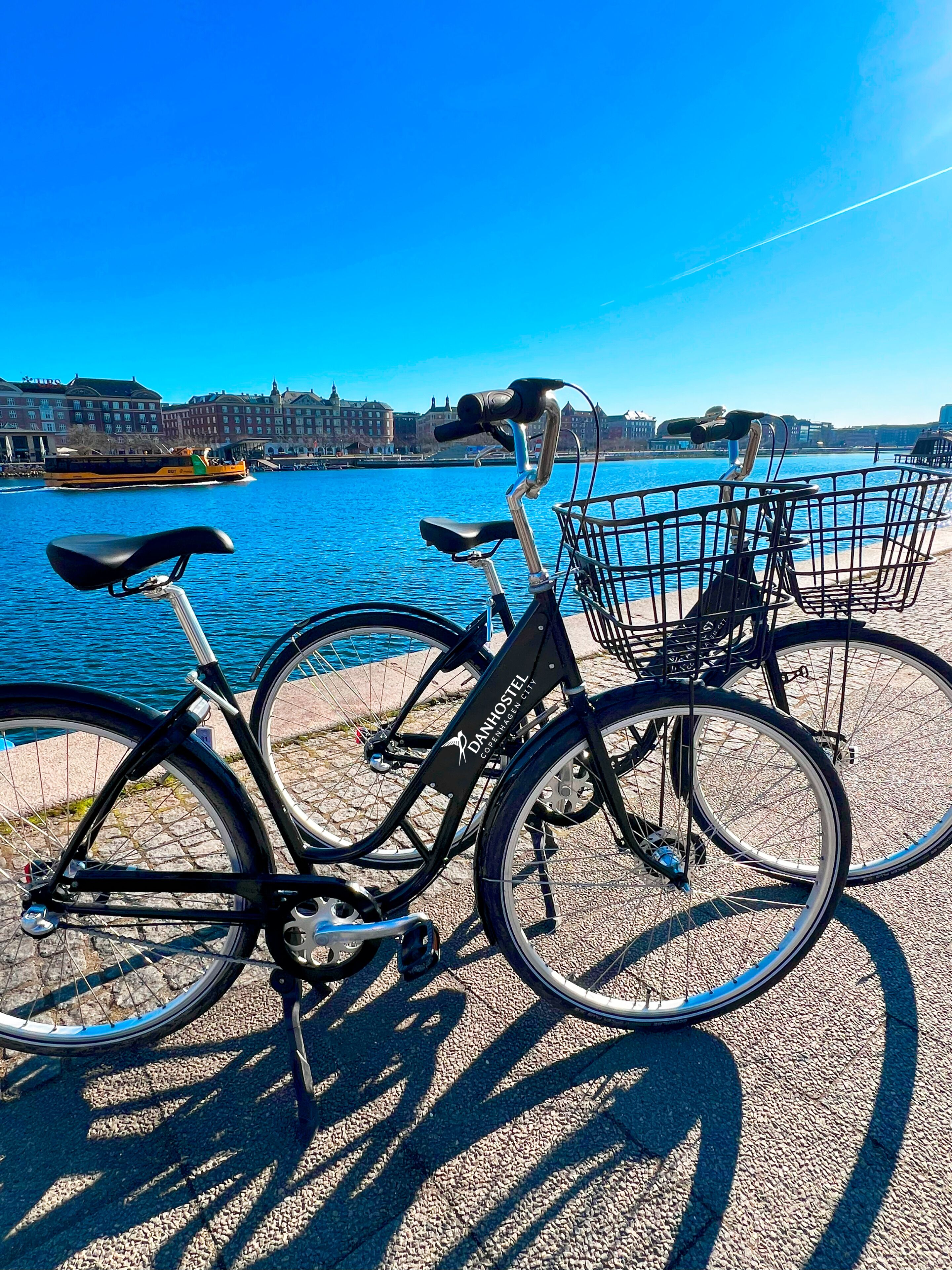 Two bicycles parked by a waterfront with a clear view of the water and buildings in the background.