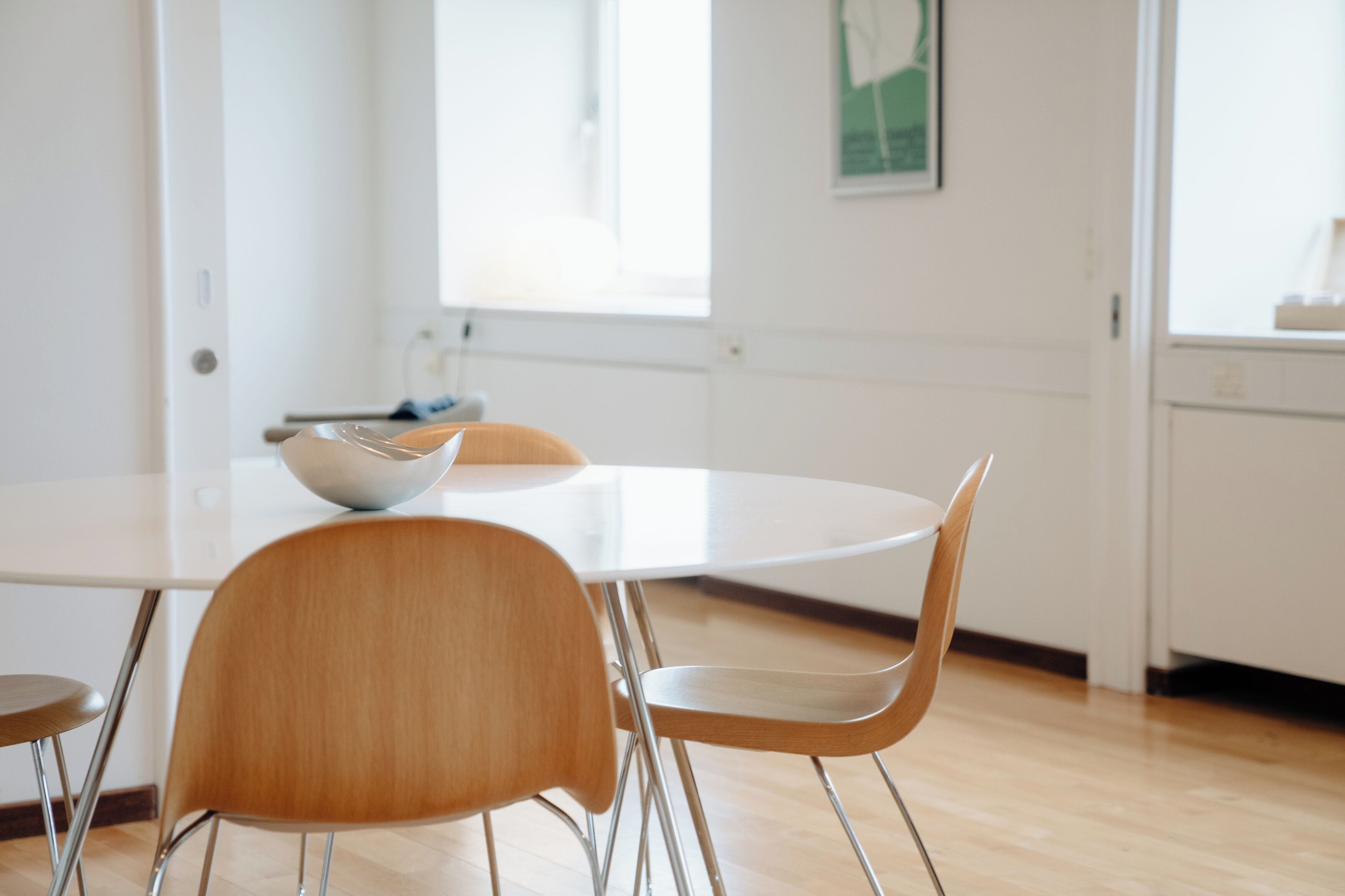 A modern dining area with a round white table, two wooden chairs, and a metal bowl on the table.