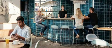 A modern bar with a blue-tiled wall, a counter with a bartender, and several patrons seated at tables.