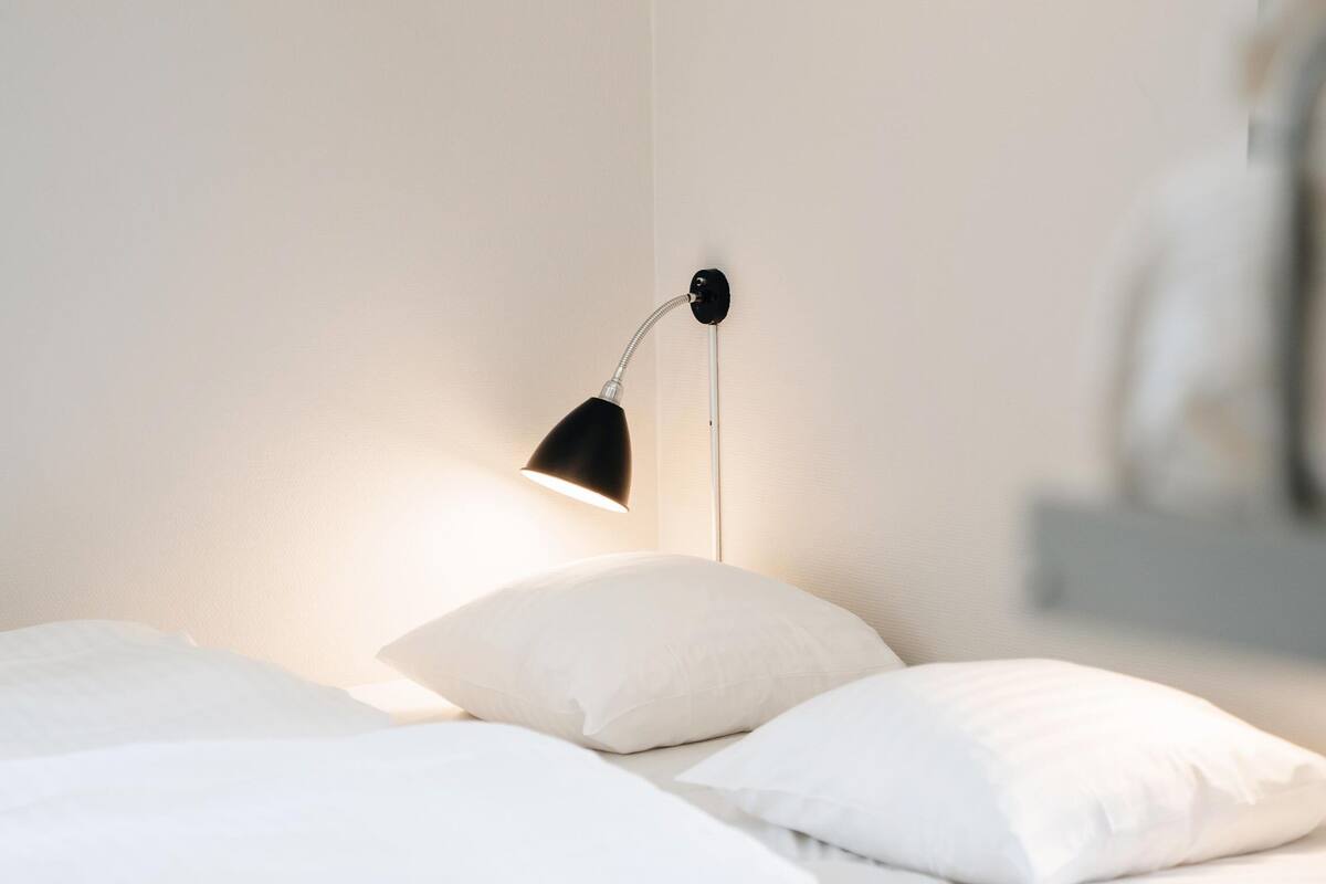 Un lit bien fait, avec du linge de lit blanc et des oreillers, une lampe noire fixée au mur et une vue partielle d’une armoire blanche.