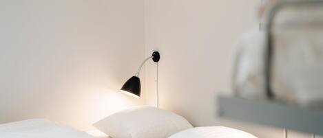 Un letto rifatto con lenzuola e cuscini bianchi, una lampada nera a parete e una parziale vista di un armadio bianco.