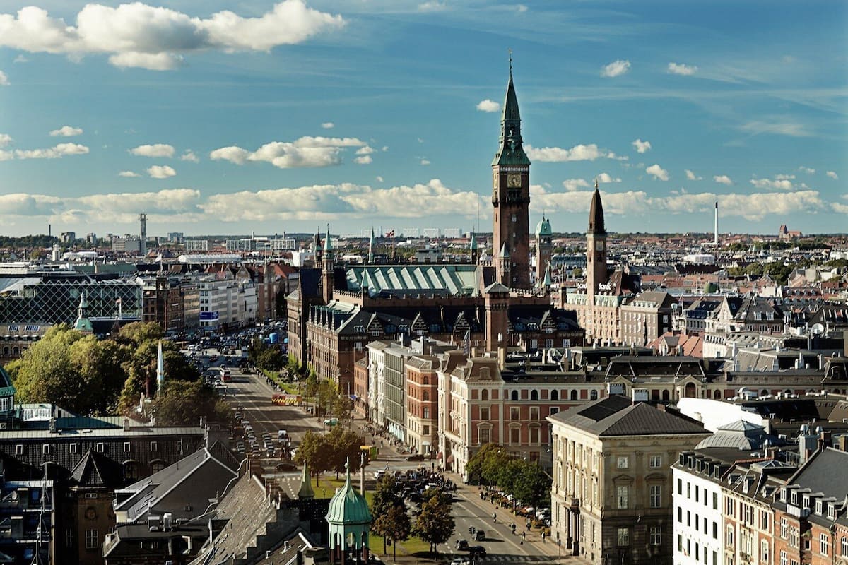 Un paysage urbain historique, avec des bâtiments remarquables, notamment un beffroi et une église surmontée d’un toit végétalisé.