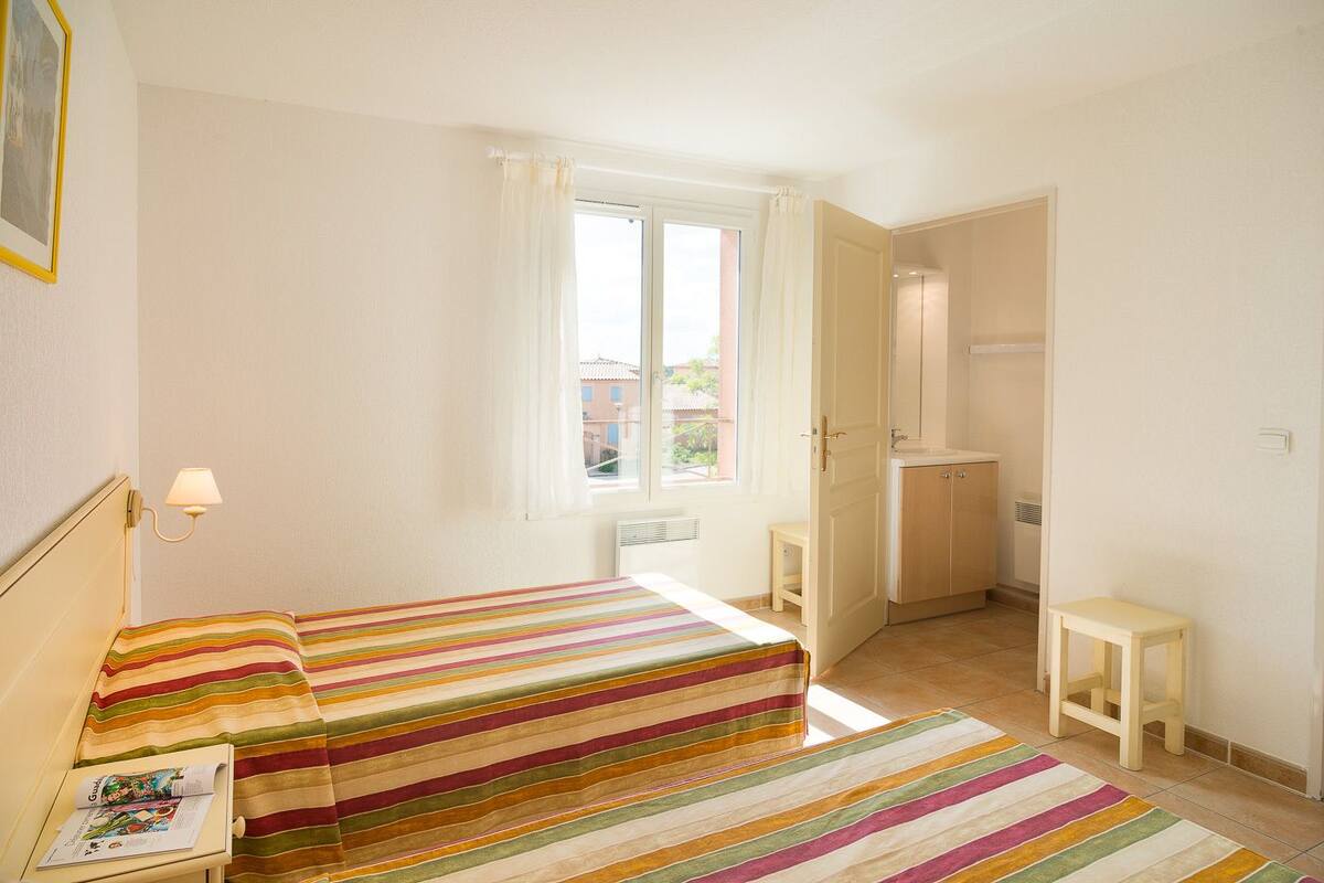 Une chambre à coucher comprenant un lit, une table de chevet, une lampe, une fenêtre avec des rideaux, une salle de bain avec un lavabo et des placards, et un petit tabouret.