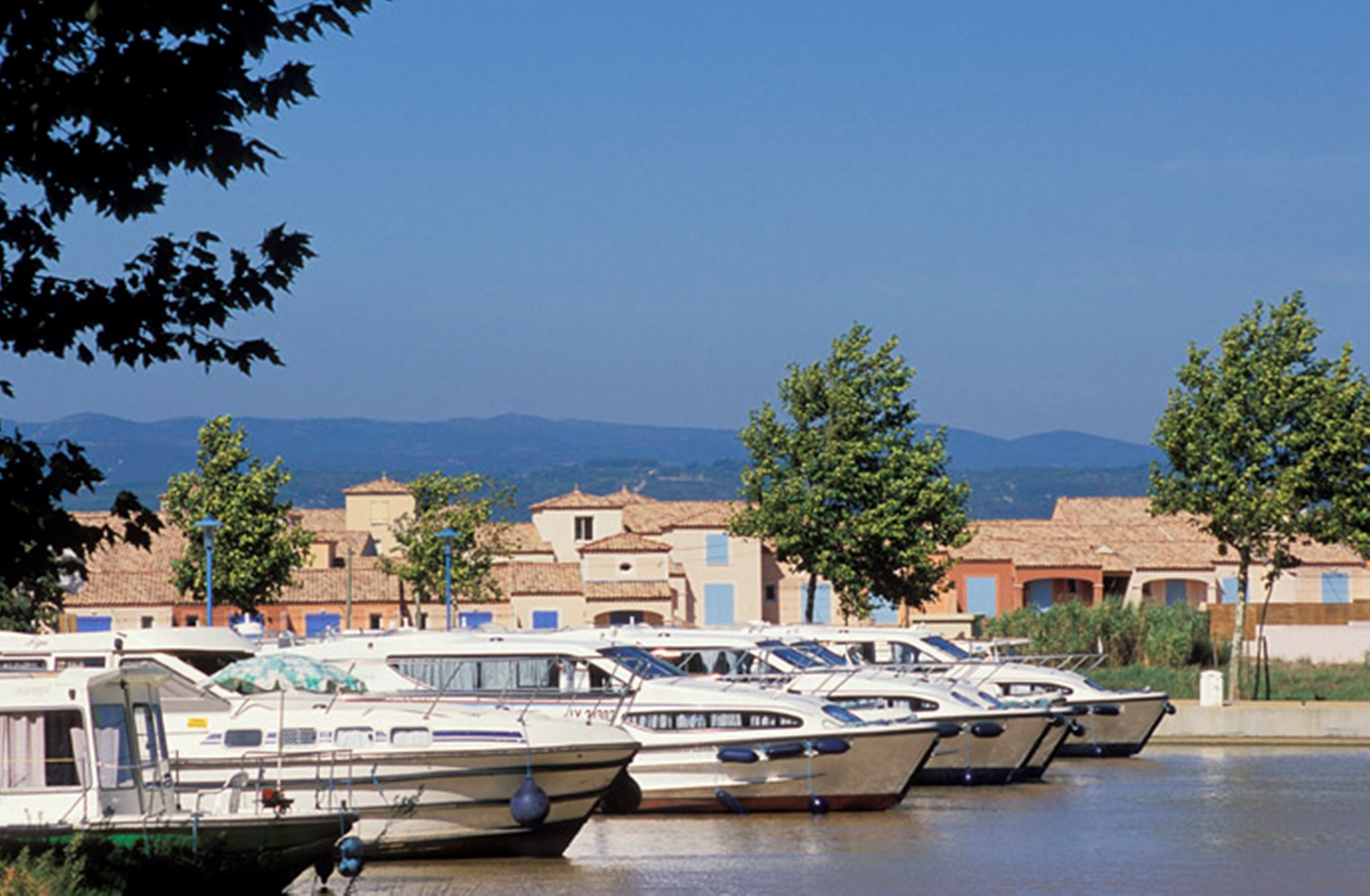 Marina com vários barcos atracados, cercada por edifícios com arquitetura mediterrânea e árvores.