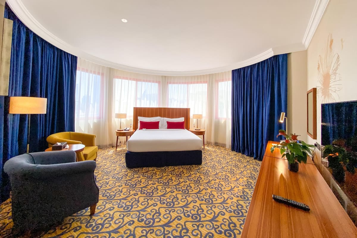 Habitación de hotel con una cama grande, dos mesitas de noche, un escritorio con una planta, una silla y un ventanal con cortinas.