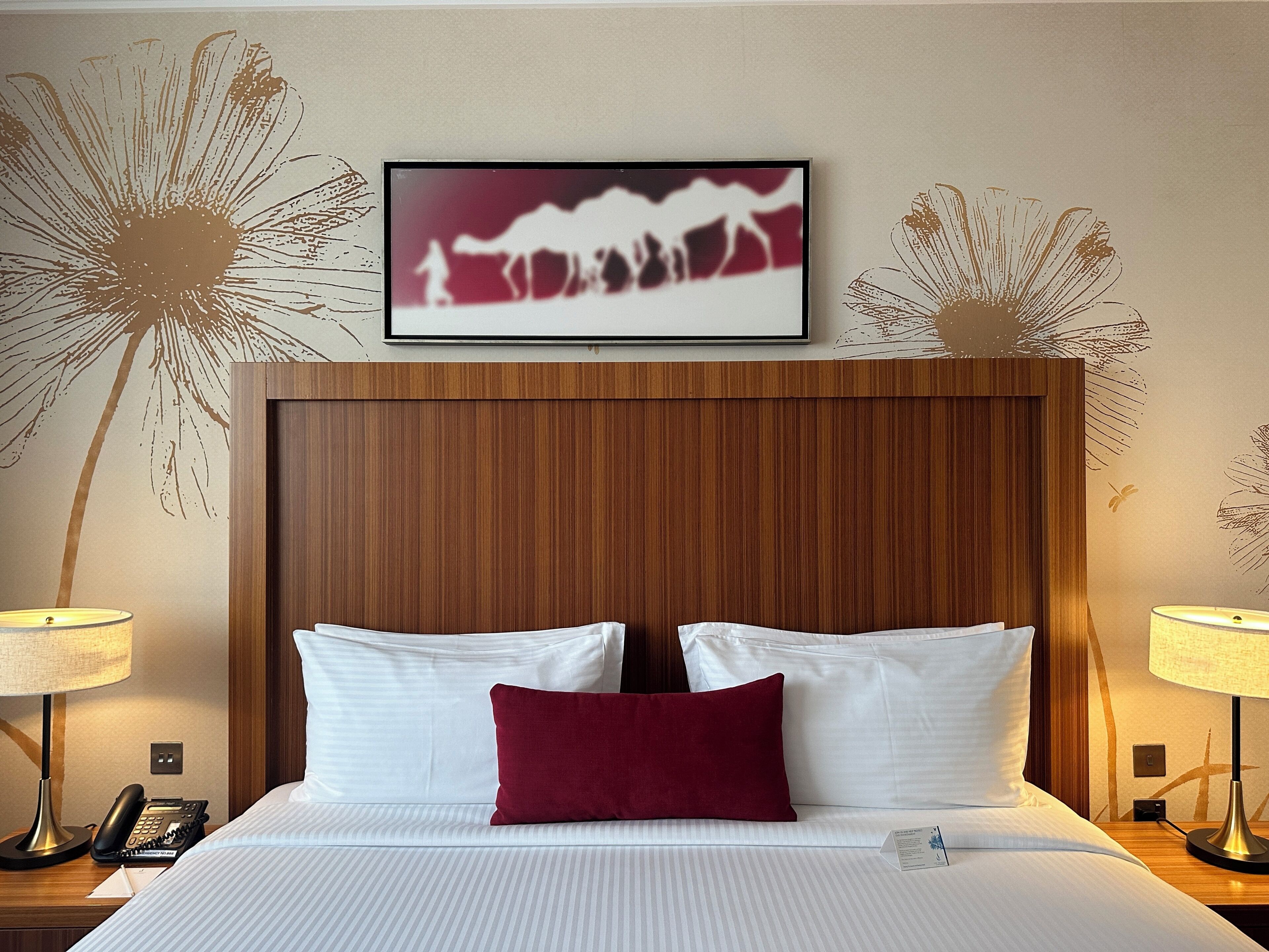 A hotel room with a wooden headboard, white bedding, a red accent pillow, and a framed picture above the bed.
