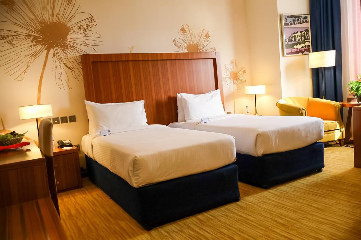 Habitación de hotel con dos camas, cabeceros de madera, mesitas de noche con lámparas, una mesita pequeña con un bol rojo y una silla amarilla.