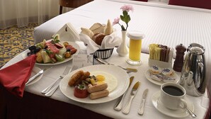 Una habitación de hotel con una cama bien tendida, una bandeja de desayuno con huevos, salchichas y tostadas, una taza de café y una fuente de fruta.