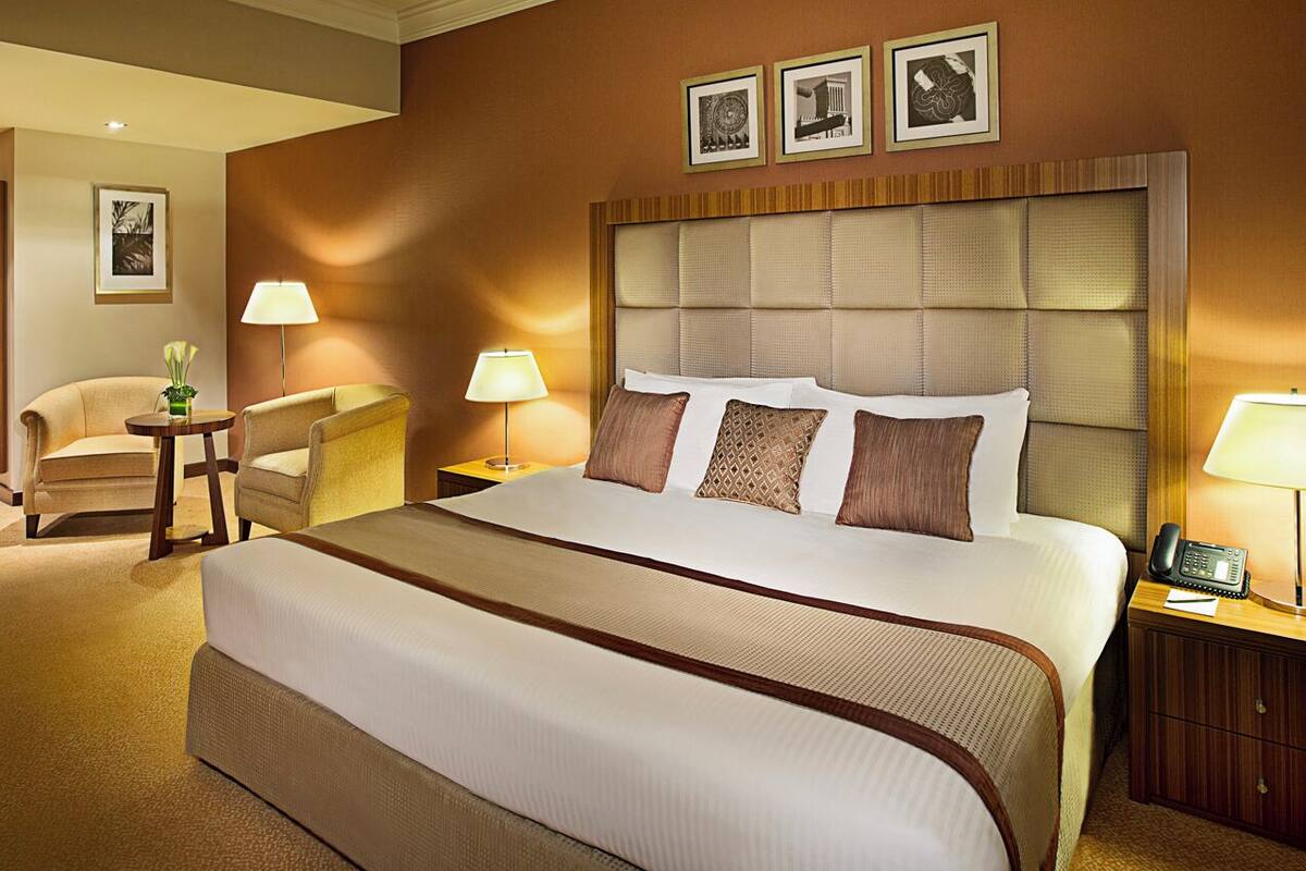 Habitación de hotel con una cama grande, dos mesitas de noche con lámparas, una silla, un escritorio con silla y cuadros en la pared.