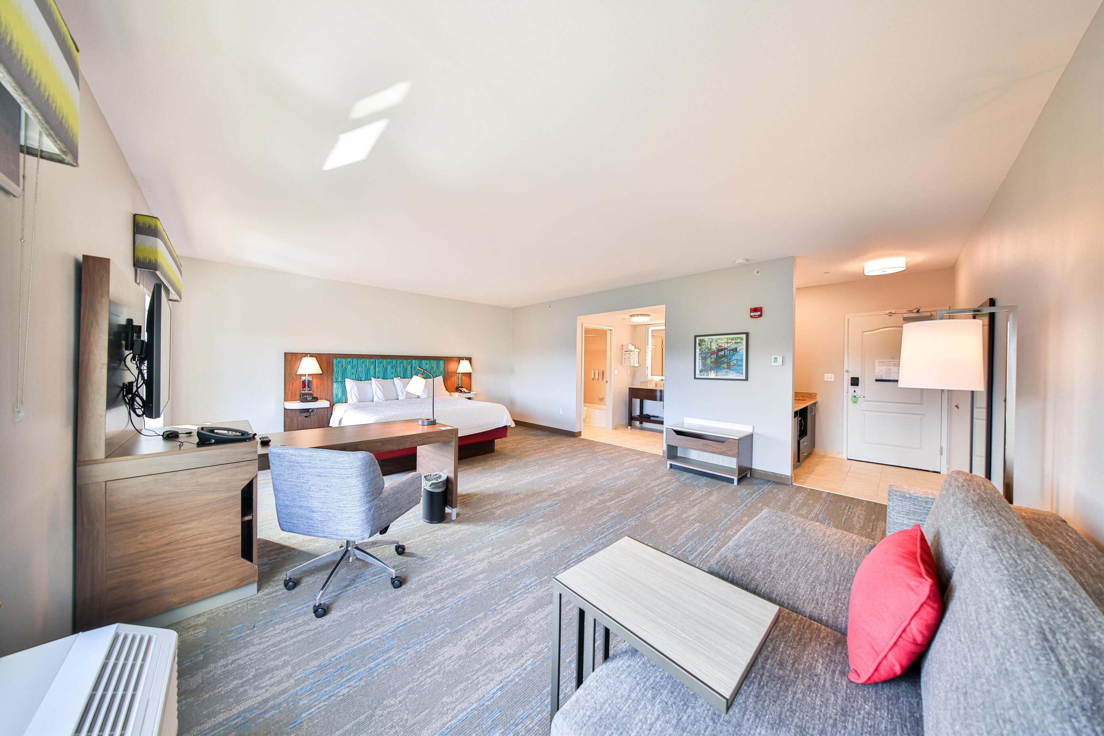 A hotel room with a bed, desk, chair, and a sofa with a red pillow.