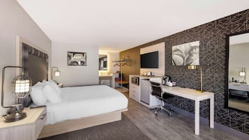 A modern hotel room with a bed, desk, and chair, a small kitchenette, and a large mirror.