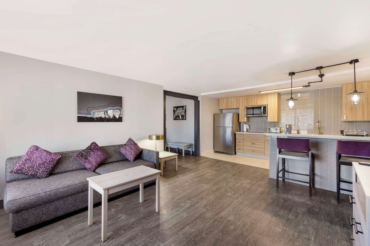 A modern living room with a grey sofa, a small table, and a kitchen area with wooden cabinets and stainless steel appliances.