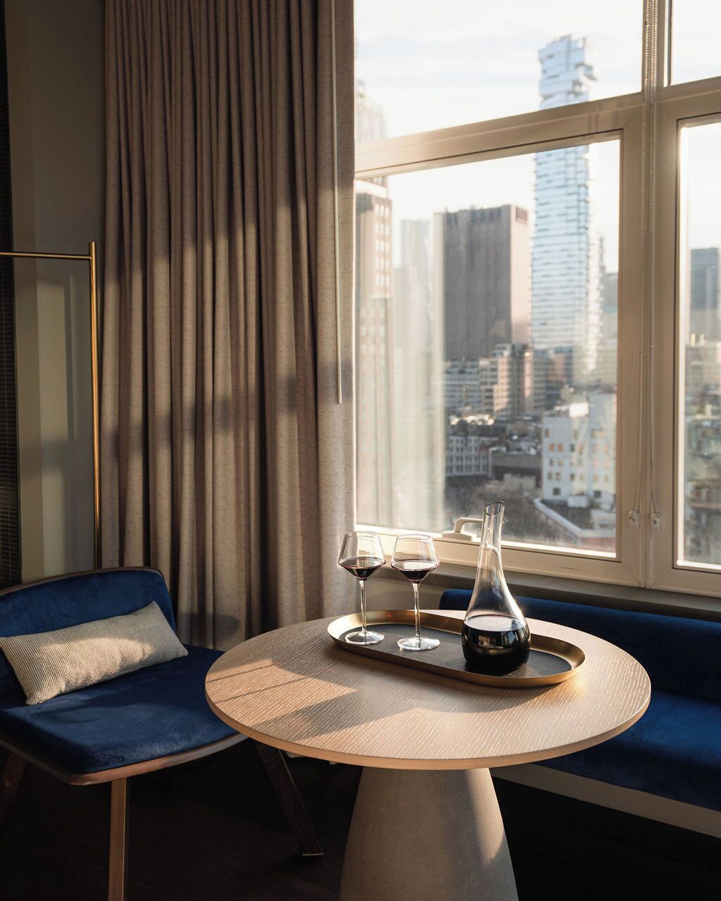 Una habitación de hotel moderna con vista a la ciudad, una mesa redonda con una bandeja, dos copas de vino y una vela.
