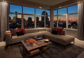 A modern living room with a large sectional sofa, a coffee table with books and flowers, and a city view through large windows. - ModernHaus SoHo (New York)