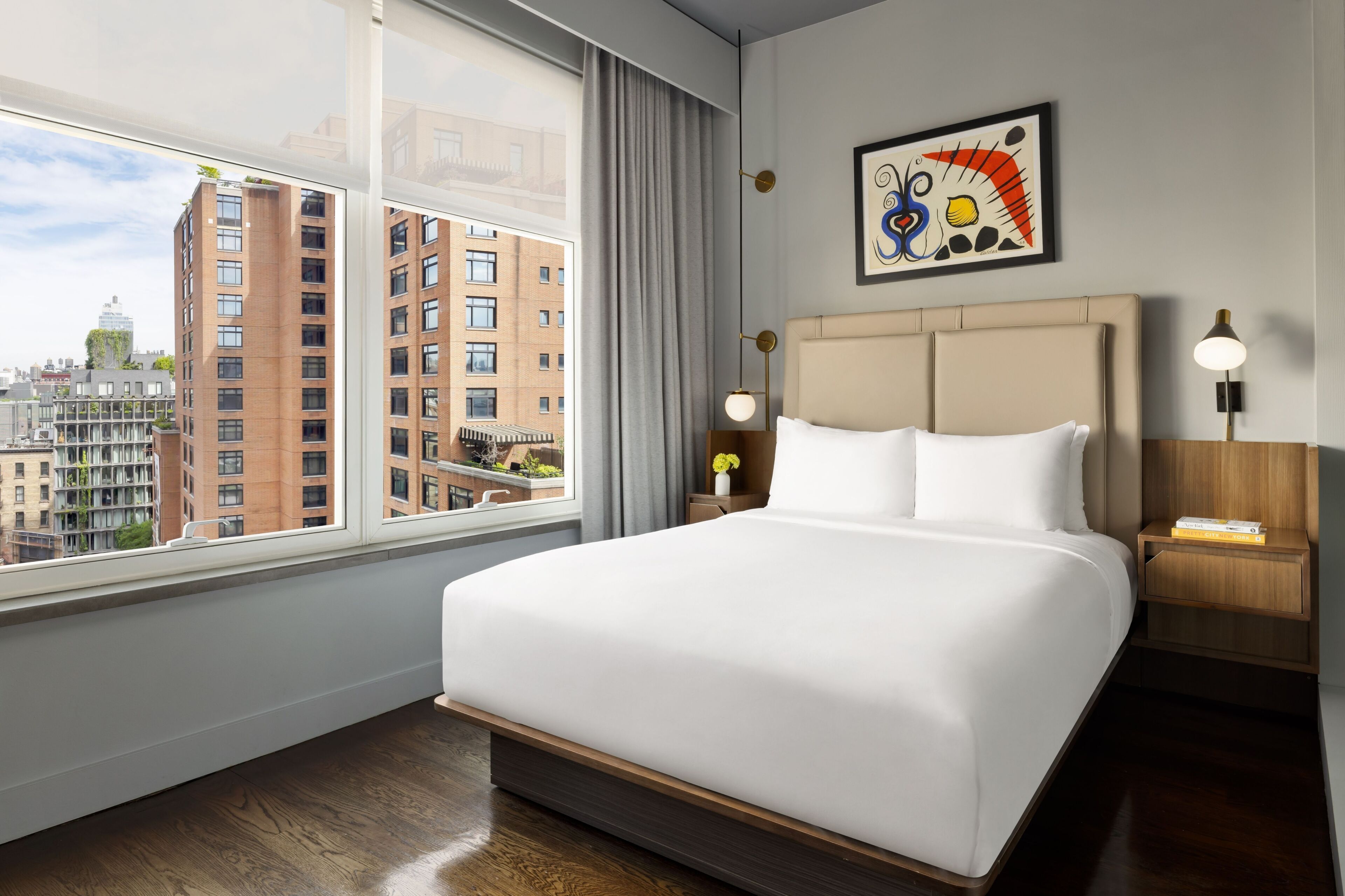 A hotel room with a large bed, a nightstand, a lamp, a painting, and a view of city buildings.