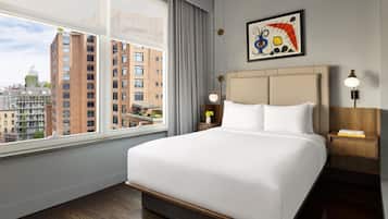 A hotel room with a large bed, a nightstand, a lamp, a painting, and a view of city buildings.
