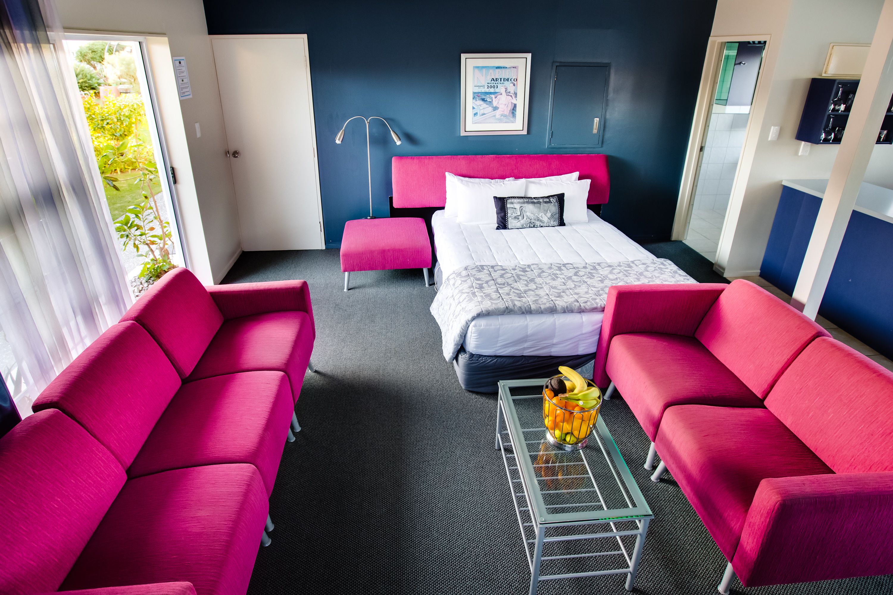 A hotel room with a bed, two red sofas, a small table with fruit, and a view of greenery outside.
